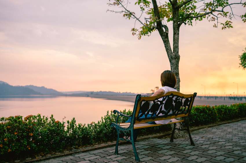 Rear-view-mature-woman-looking-lake-while-sitting-bench-against-sky-sunset - travel and tour world