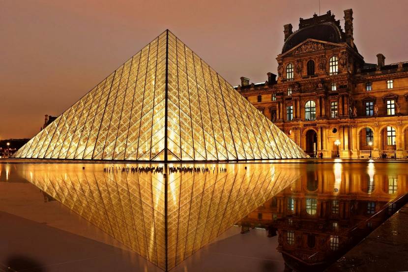 A wide-angle view of the iconic glass pyramid entrance of the Louvre Museum in Paris under a clear sky, currently affected by temporary staff closures.