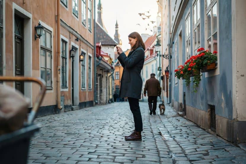Woman in a dark coat stands on a cobblestone street, taking a photo with her phone; pastel buildings and flower boxes line the alley.