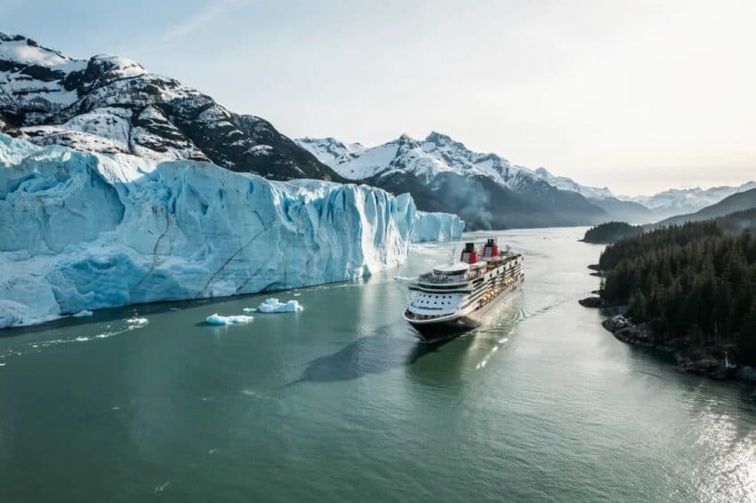 Cruise ship sails through a fjord past a towering blue glacier with snow-capped mountains in the background and evergreen trees on the right coast.