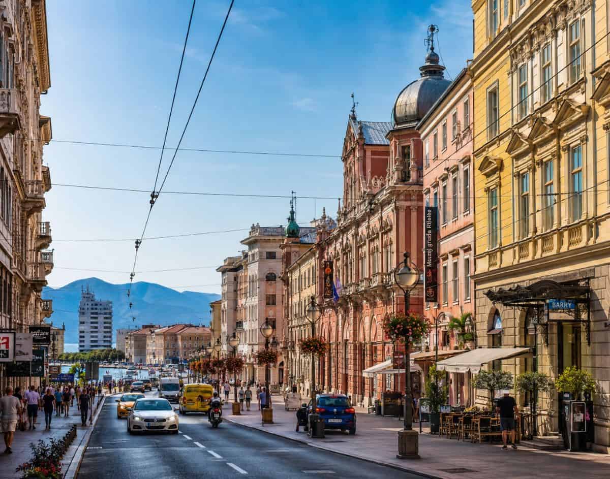 A vibrant croatian city street with historic architecture, busy traffic, caf&eacute;s, pedestrians, and scenic mountains under bright mediterranean skies.