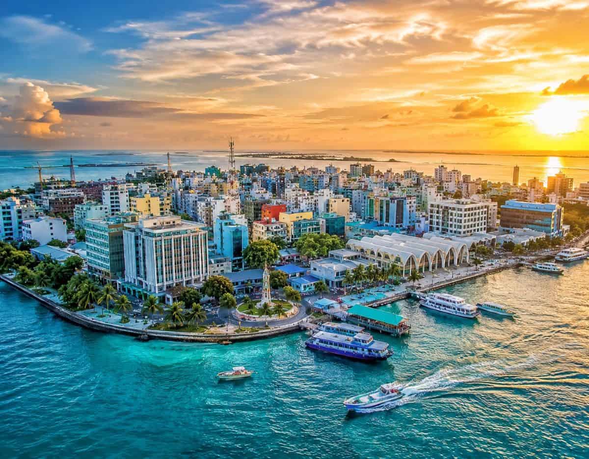Maldives cityscape glows at sunset, with turquoise waters, colourful buildings, palm-lined shores, boats, and a vibrant tropical skyline.