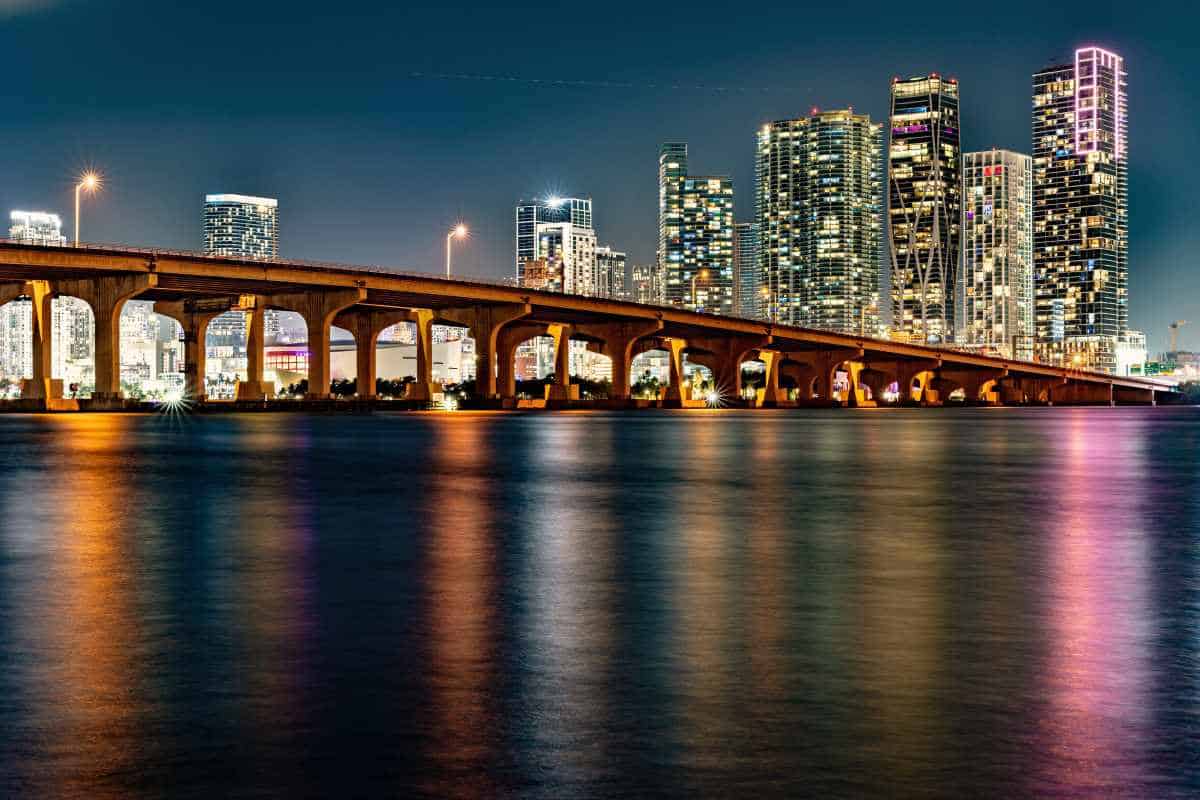 Miami - travel and tour world A twilight view of the miami skyline across the water, featuring illuminated high-rise buildings and a long highway bridge spanning the bay, with bright city lights reflecting on the water's surface.