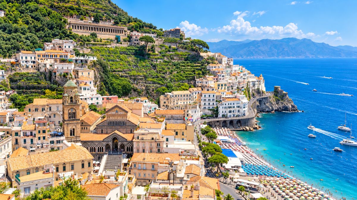 Aerial view of amalfi on italy&rsquo;s amalfi coast, showing colourful hillside buildings, terraced greenery, historic cathedral, coastal road, sandy beach and bright blue mediterranean sea