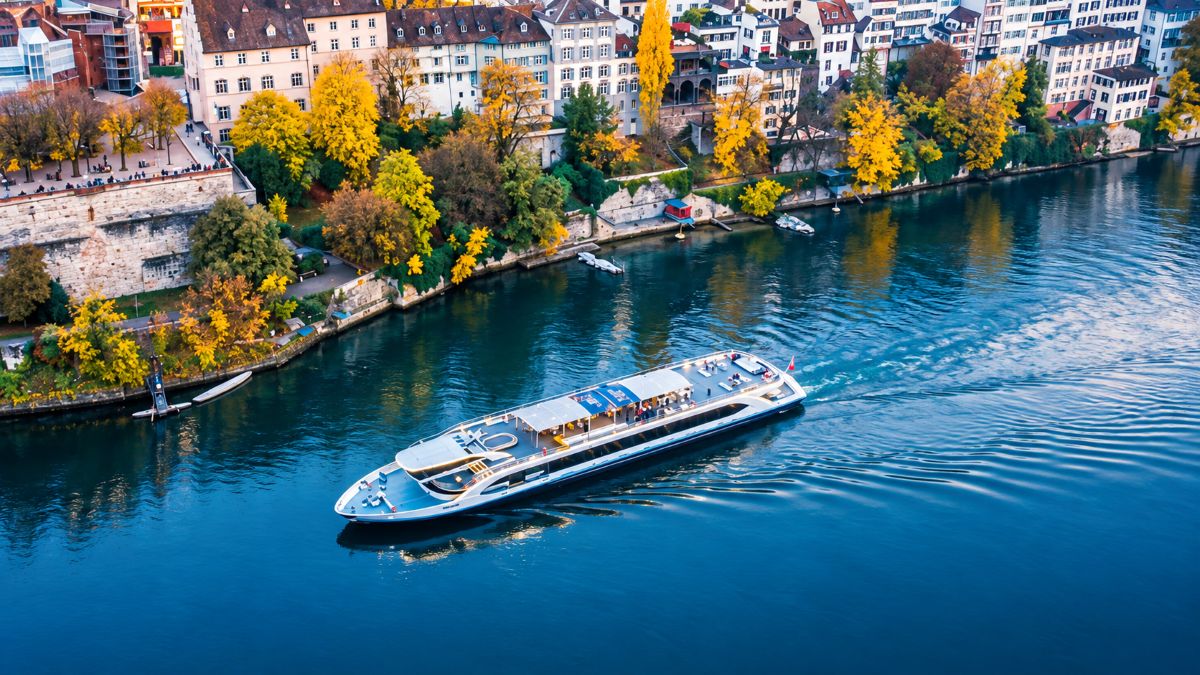 Aerial view of a modern river cruise boat sailing along a blue river beside a historic european city lined with golden autumn trees