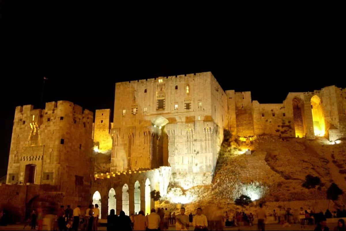 Night view of the illuminated aleppo citadel in syria, showcasing its ancient stone architecture and defensive walls lit up against a dark sky, with visitors gathered below.
