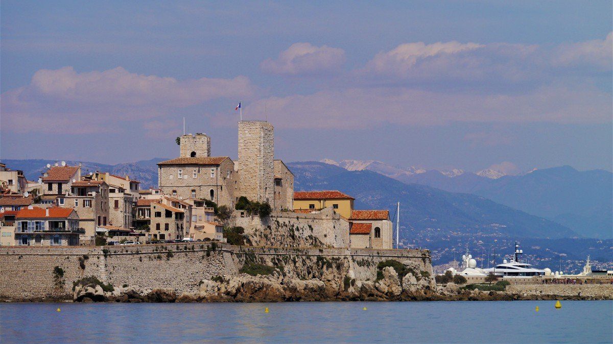 A wide shot of the coastal town of antibes, france, featuring historic stone ramparts, traditional terracotta-roofed buildings, and a mediaeval fortress castle overlooking the blue sea, with distant hazy mountains under a cloudy sky.