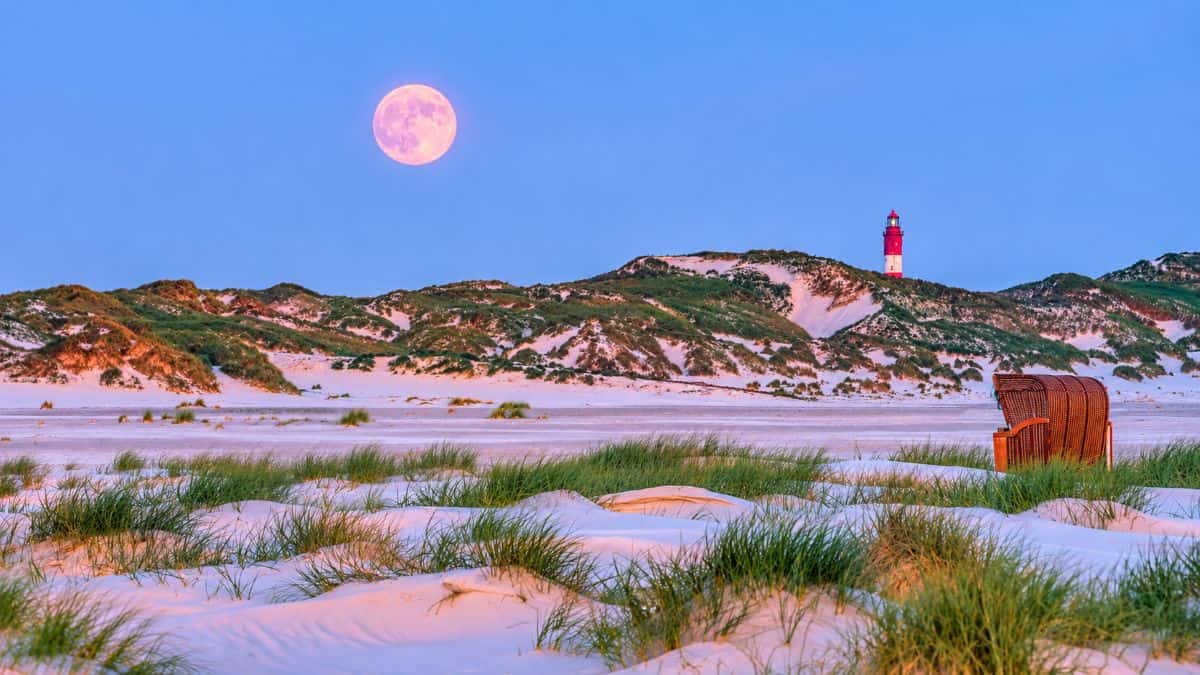 A tranquil sandy beach with grassy dunes, a red-and-white lighthouse, a wicker beach chair and a glowing pink moon under a blue evening sky.