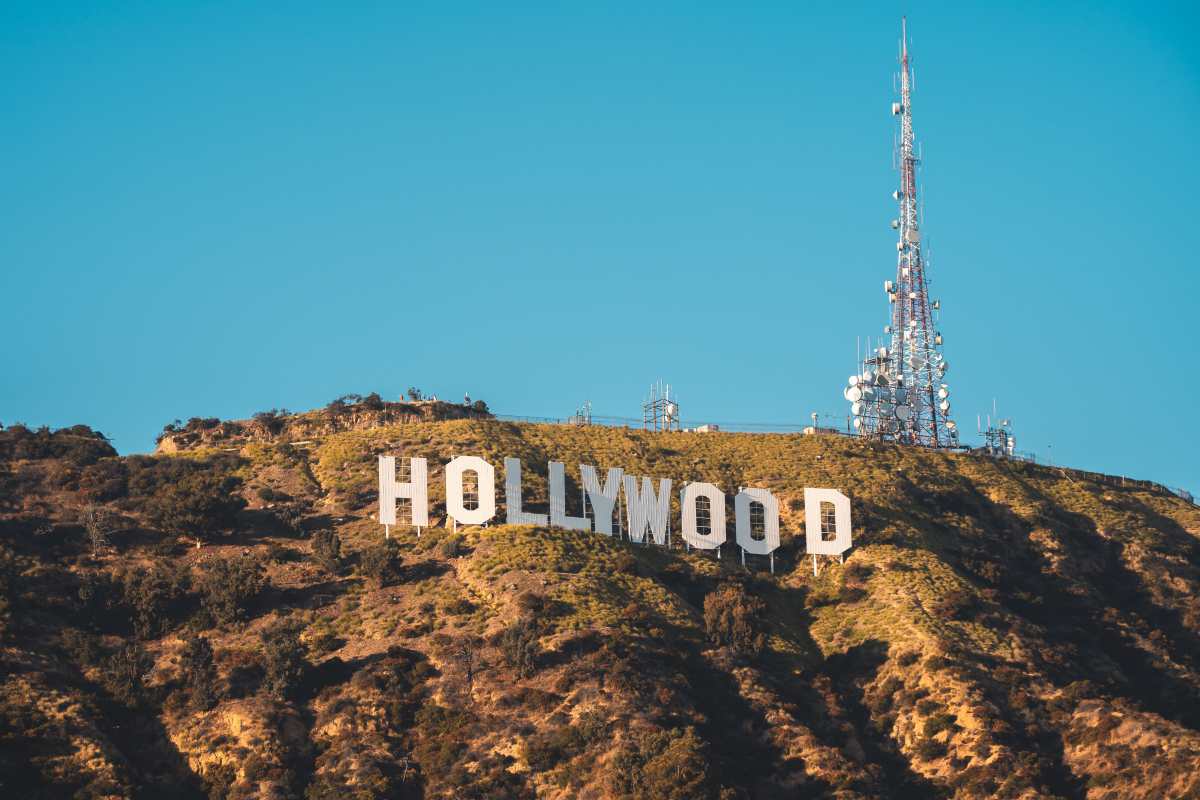 A wide-angle daytime shot of the white hollywod sign set against a dry, brush-covered hill under a clear blue sky, with a tall telecommunication tower rising behind the letters to the right.