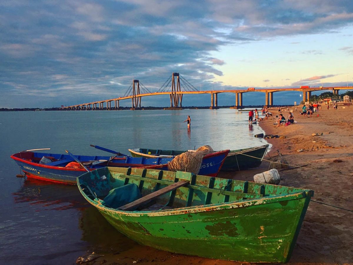Wooden fishing boats resting on a sandy riverbank at sunset, with a long suspension bridge stretching across the wide river under a cloudy sky in the background.