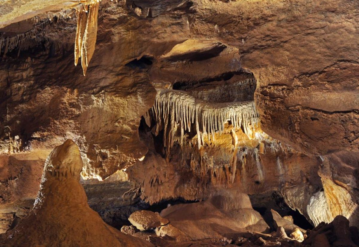 Interior view of a czech show cave showing intricate rock formations, hanging stalactites, and ground stalagmites illuminated in a warm glow.