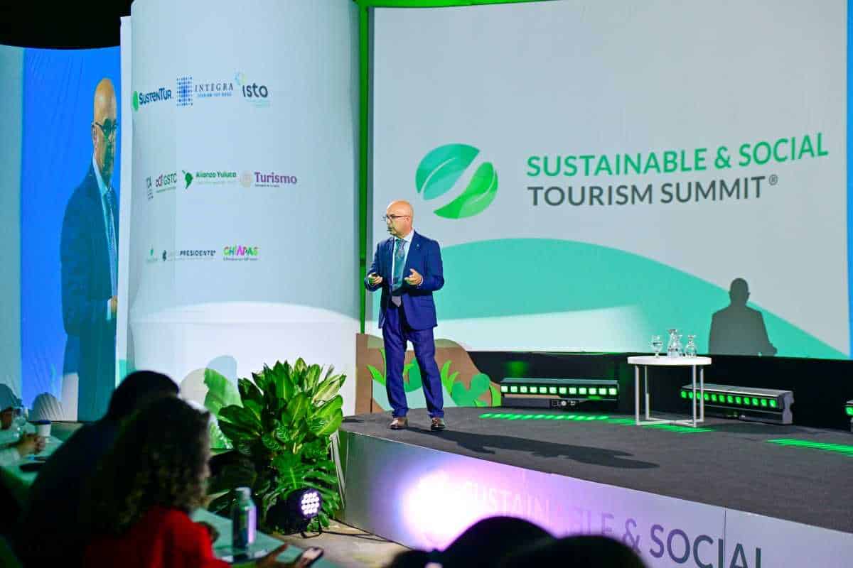 El salvador - travel and tour world A speaker in a blue suit stands on a stage presenting at the sustainable & social tourism summit, with a large screen in the background displaying the event's logo and various partner emblems.