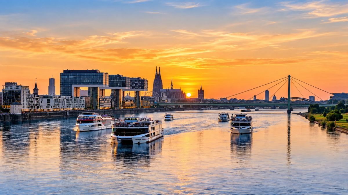 Golden-hour river view of cologne with sightseeing boats, modern waterfront buildings, historic twin-spired cologne cathedral and bridges reflected on the rhine.