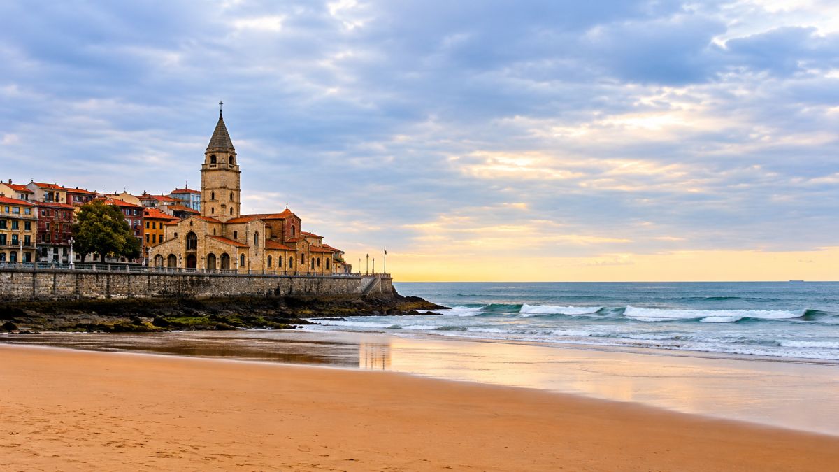 Historic seaside church and coastal town beside a wide sandy beach with ocean waves and a dramatic cloudy sunset sky