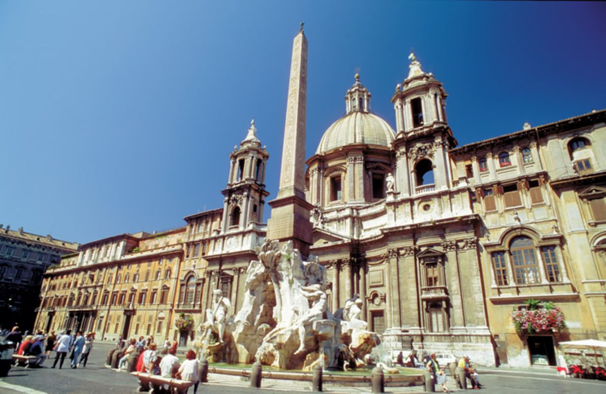 A sunlit daytime view of piazza navona in rome, italy, featuring the historic fountain of the four rivers with its central obelisk, flanked by ornate classical architecture and the church of sant'agnese in agone under a clear blue sky.