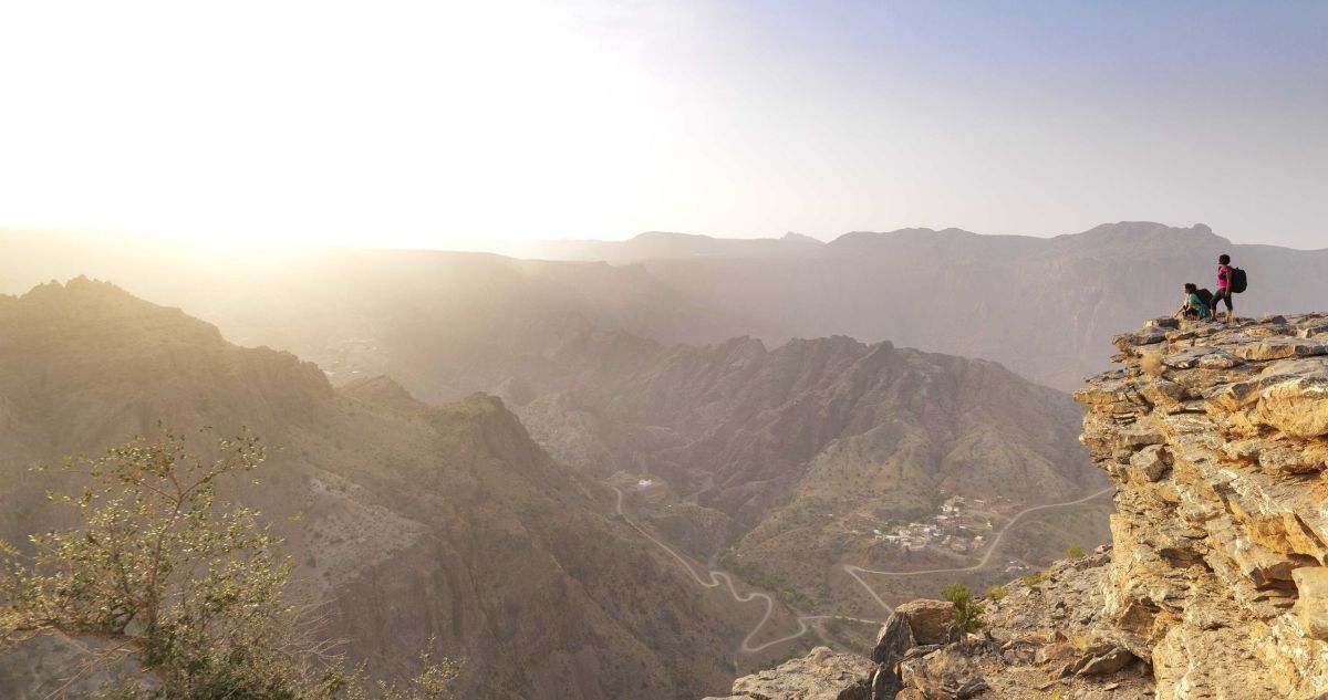 Two hikers stand on a rocky cliff edge overlooking a vast, rugged canyon landscape with winding mountain roads and a hazy sunset sky.