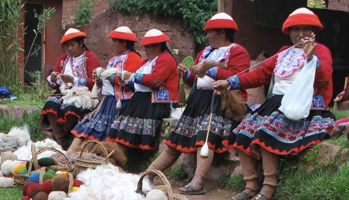 Five women in traditional red-and-white hats and patterned skirts sitting outdoors, spinning yarn and working with wool in a community tourism setting.