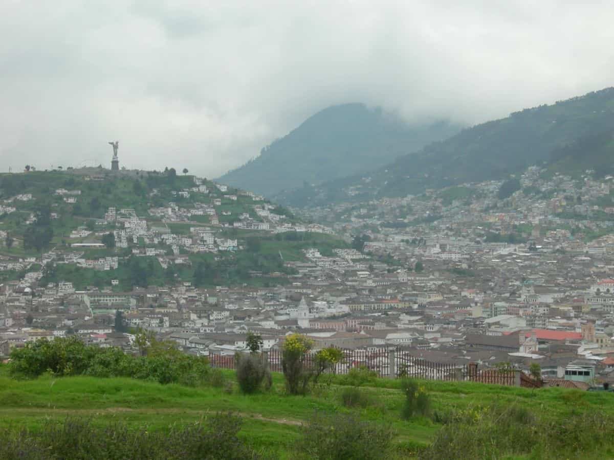 An elevated, wide-angle view of quito, ecuador, stretching across a valley and climbing up the surrounding green andean hills under an overcast sky. In the foreground, a grassy hillside with a fence looks out over the dense city rooftops and historic architecture, while a prominent statue sits atop the distant hill on the left.