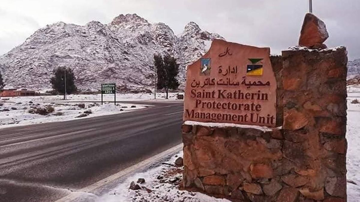 A stone sign reading "saint katherin protectorate management unit" in english and arabic sits beside an asphalt road, with snow-covered mountains and ground in the background.