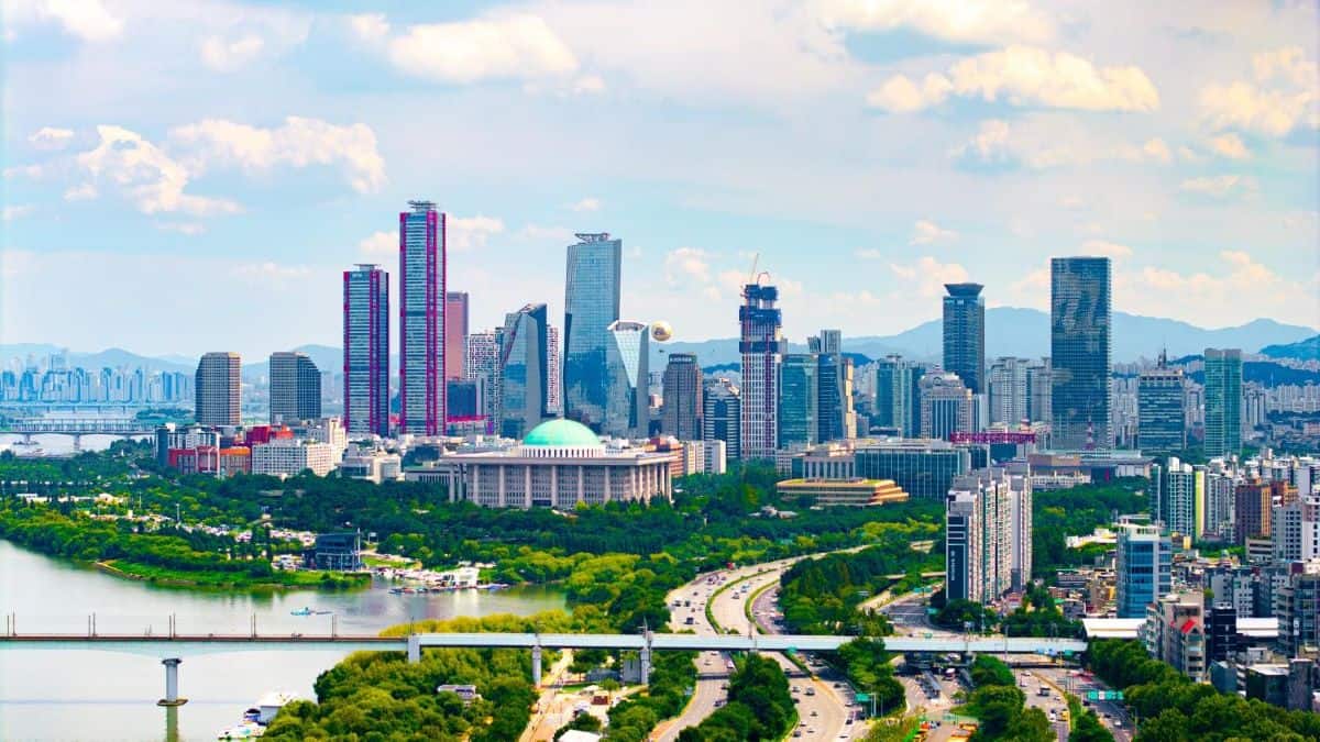 Seoul - travel and tour world An aerial view of the seoul skyline featuring modern high-rise skyscrapers, the iconic blue-domed national assembly building, lush green trees, a wide river with bridges, and a partly cloudy blue sky.