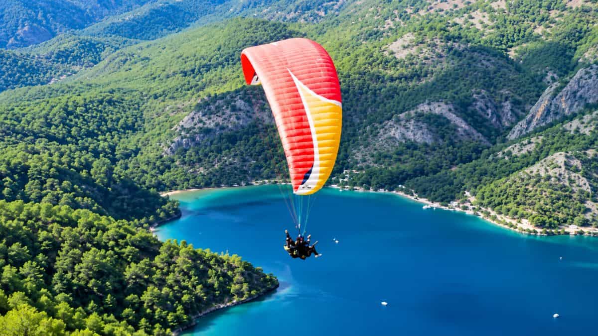 Tandem paraglider flying above a vivid turquoise mountain lake surrounded by lush green forested hills and rocky cliffs - travel and tour world Tandem paraglider flying above a vivid turquoise mountain lake surrounded by lush green forested hills and rocky cliffs.