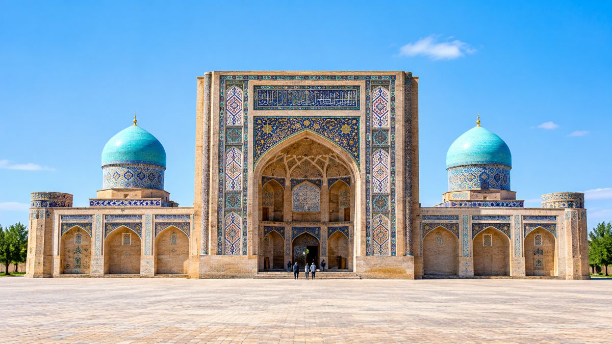 Ultra-realistic view of an ornate historic islamic madrasa with intricate blue tilework, a grand arched entrance, twin turquoise domes, and a spacious courtyard beneath a clear blue sky.