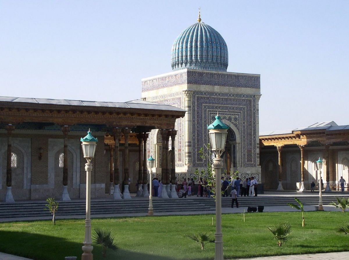 A daytime outdoor view of the imam al-bukhari memorial complex in uzbekistan, featuring a grand structure with an intricate blue and turquoise ribbed dome, geometric tilework, and a covered courtyard supported by carved wooden columns, with visitors walking on the stone pathways and lawn in the foreground.