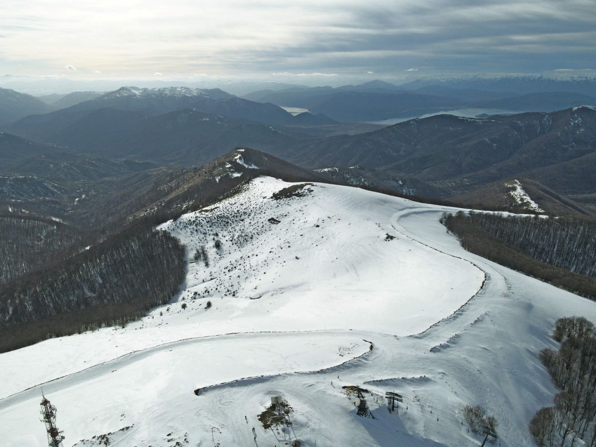 An aerial view of a snow-covered mountain ridge and ski slope in west macedonia, featuring winding tracks, sparse trees, and distant hazy mountain ranges under a cloudy sky.