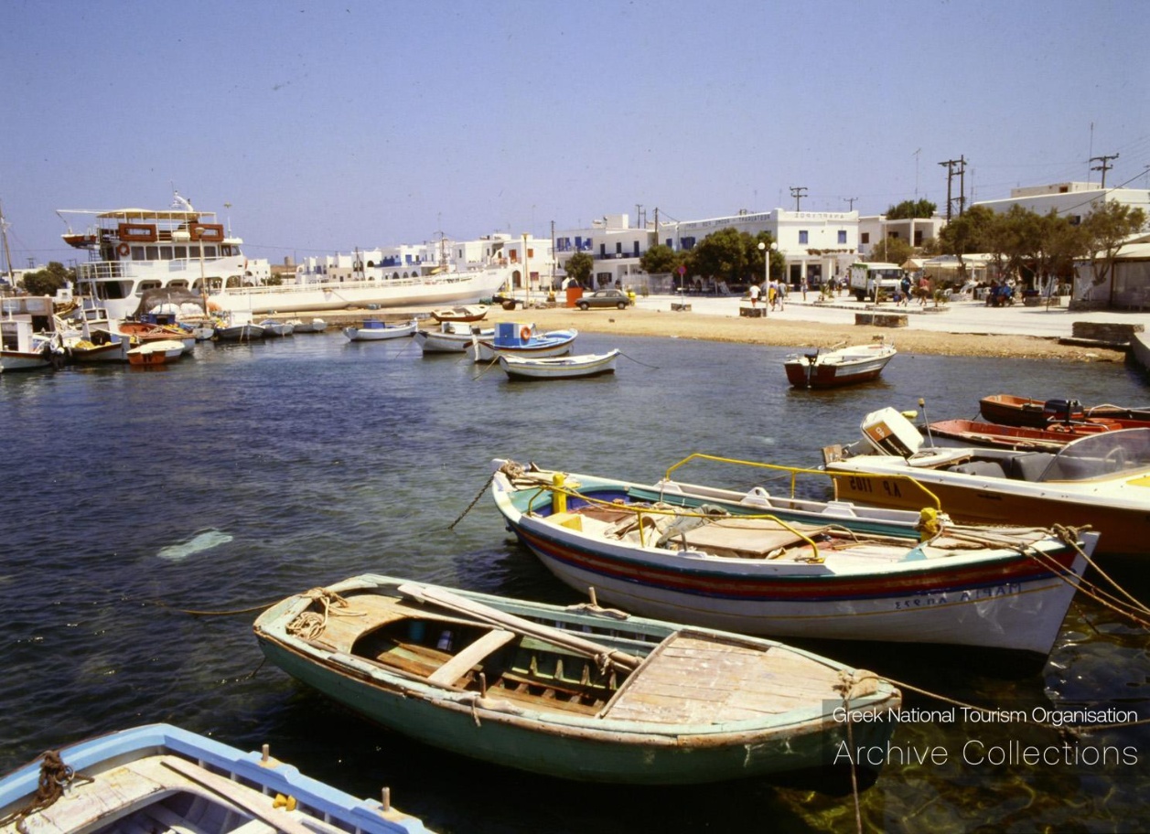 Small boats and a passenger ferry in the harbour at antiparos, greece, with white waterfront buildings behind them.