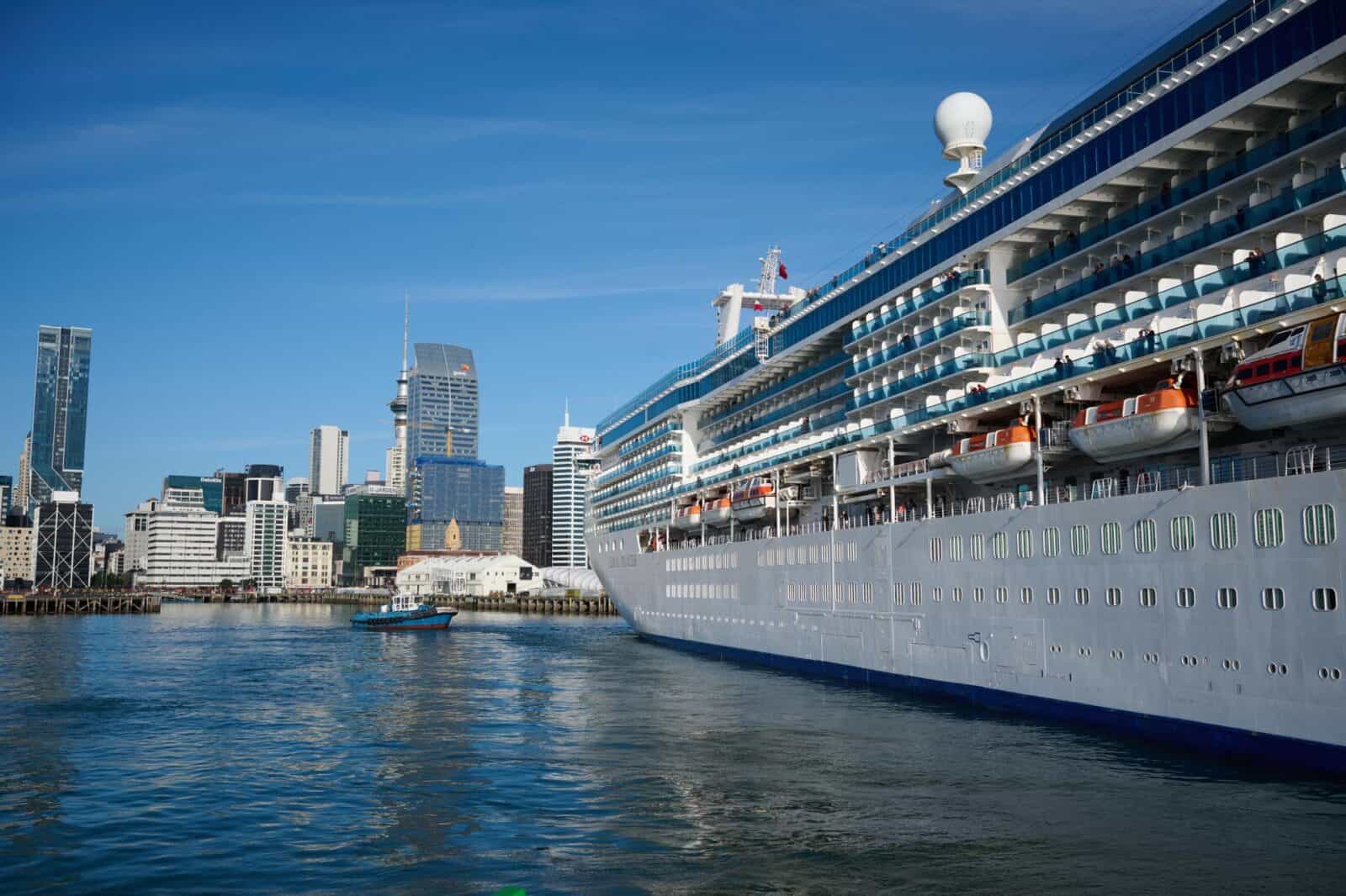 New zealand cruise tourism - travel and tour world Cruise ship beside auckland’s waterfront, with a tugboat in the harbour and the sky tower in the background.