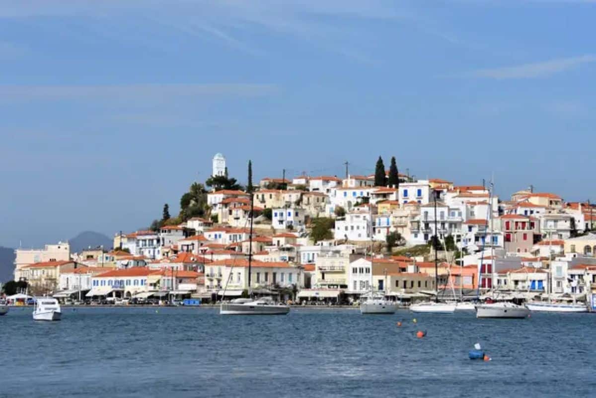 A scenic waterfront view of a greek coastal town built on a hillside, featuring whitewashed buildings with terracotta roofs, blue window shutters, and a white clock tower near the peak, with sailboats anchored in the calm blue water under a clear sky.