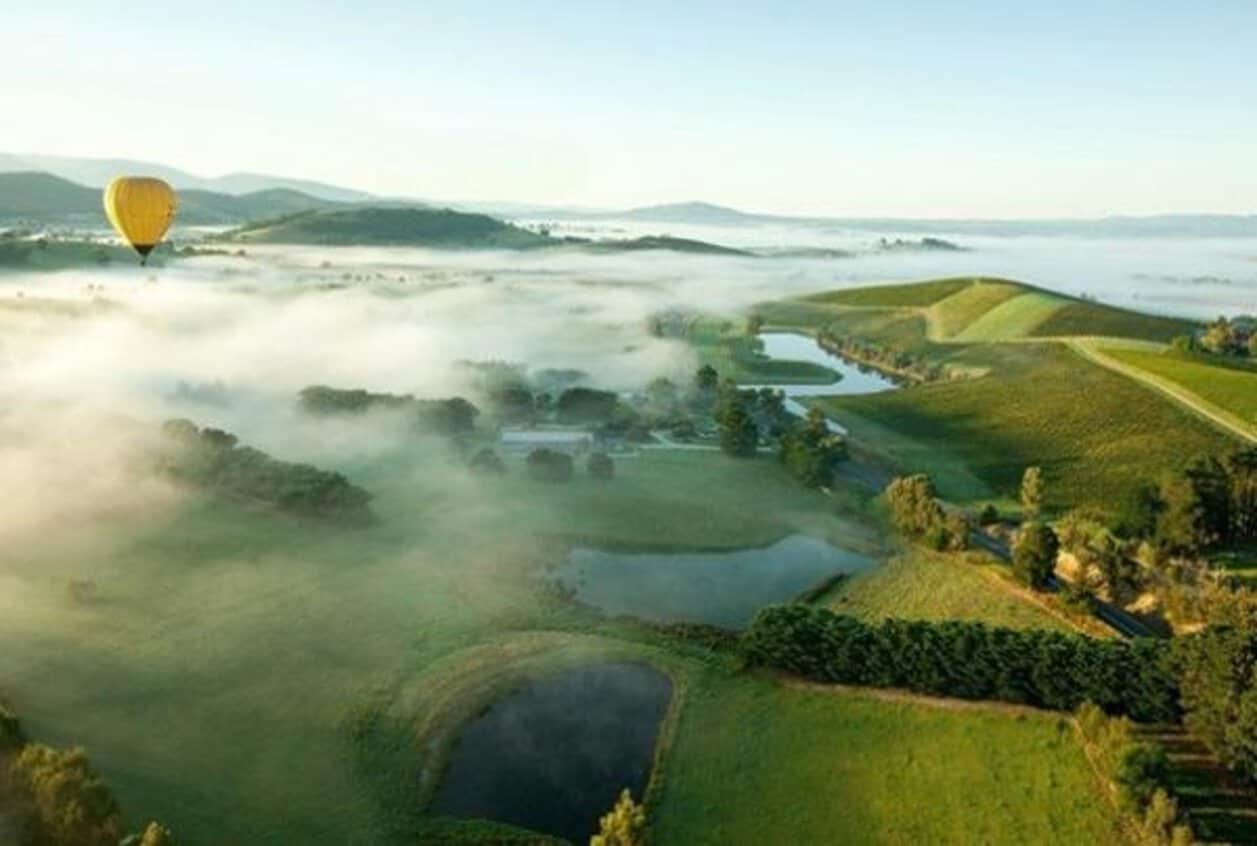 Yellow hot-air balloon above misty vineyards, rolling hills and lakes at sunrise.