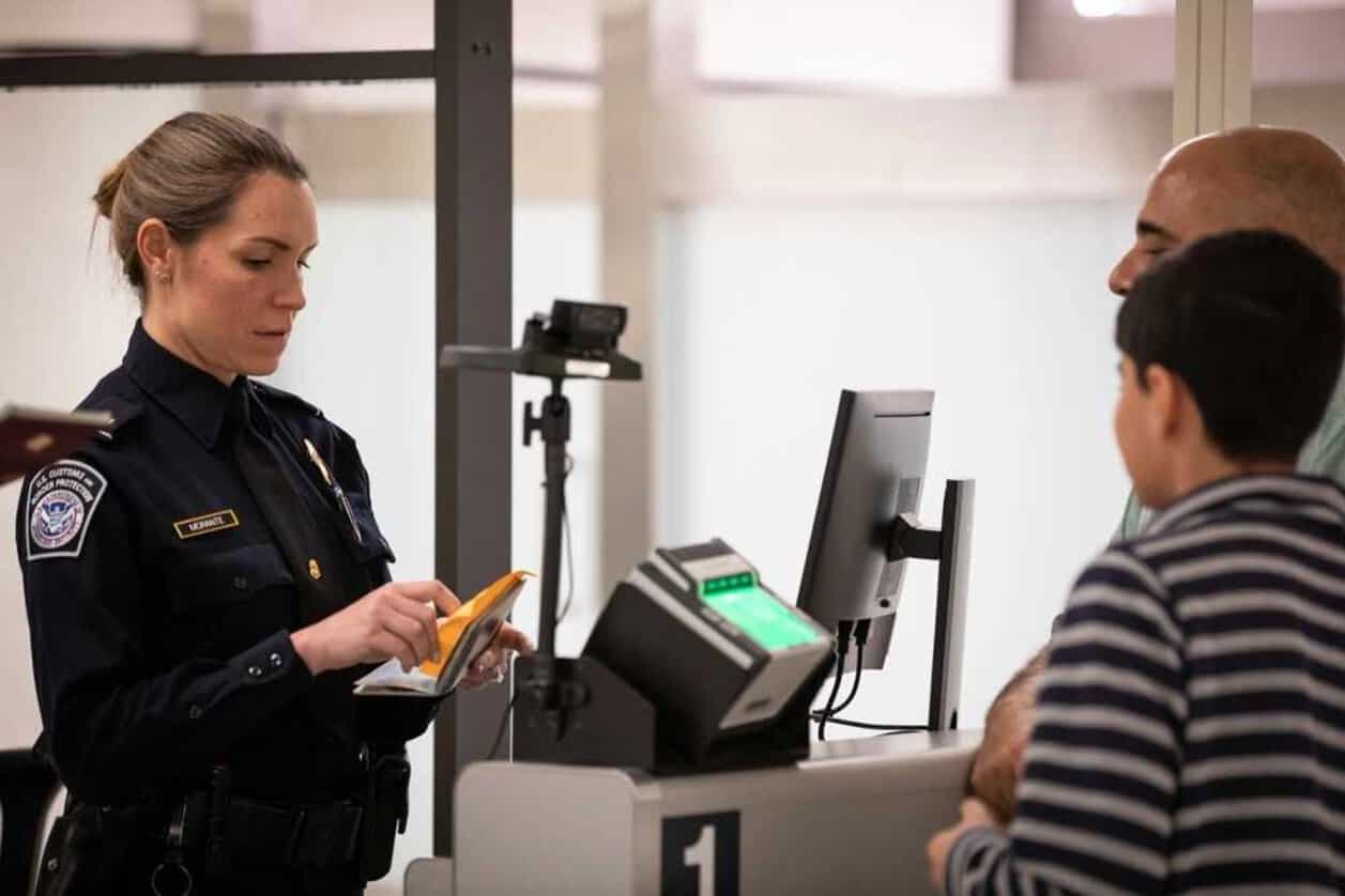 Cbp officer examining travel documents at an inspection counter while two travellers wait beside a fingerprint scanner.