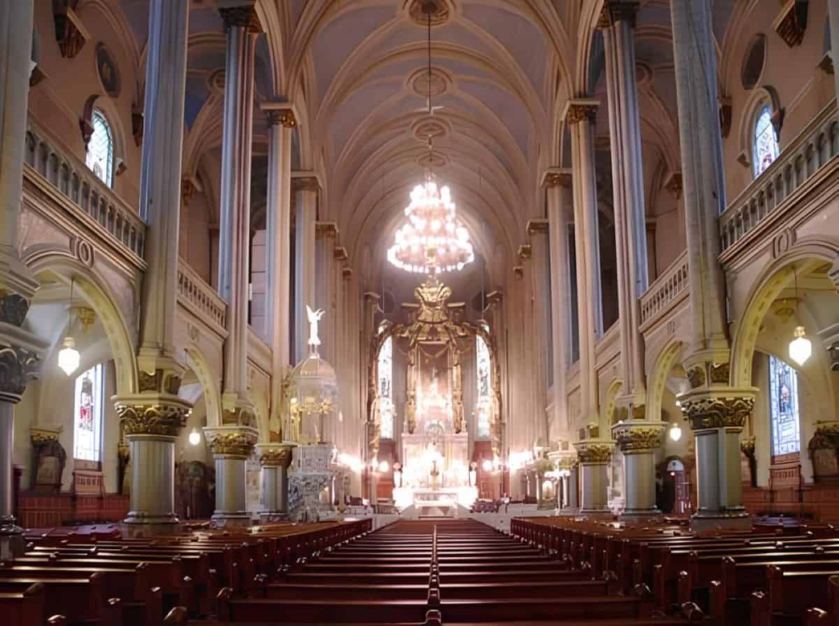 The grand interior nave of &Eacute;glise saint-jean-baptiste in quebec city, featuring wooden pews, towering columns with ornate gold capitals, vaulted ceilings, and a bright chandelier hanging over the altar.