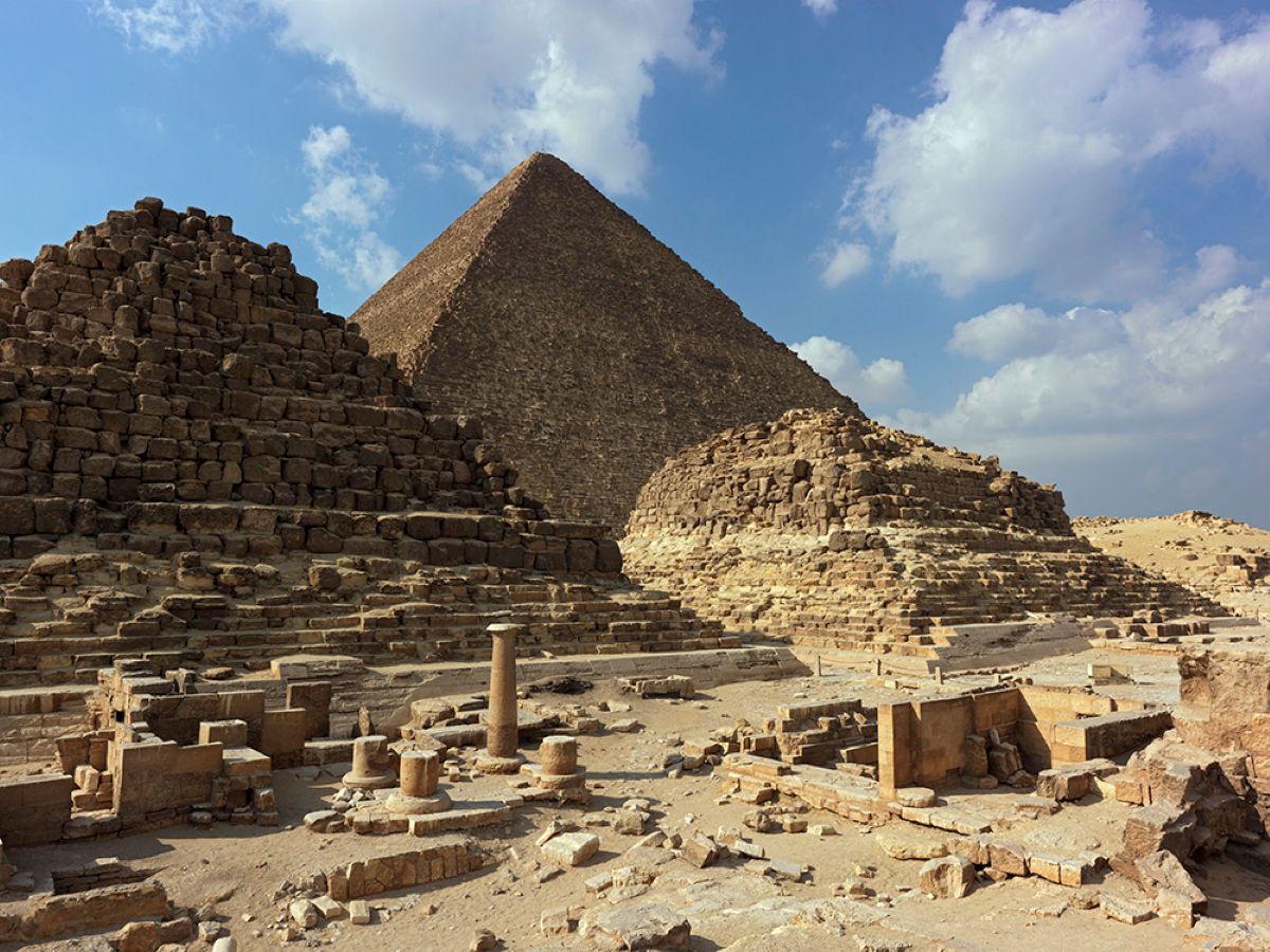 Ancient stone ruins and smaller step pyramids set against the backdrop of a large egyptian pyramid under a bright blue sky with fluffy white clouds.