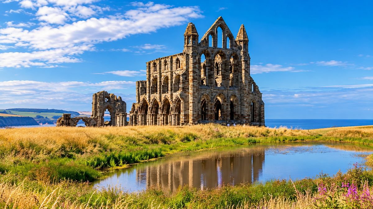 Gothic abbey ruins beside a coastal meadow and reflective pond, with golden grasses, blue sea, distant cliffs and a bright cloud-filled sky.