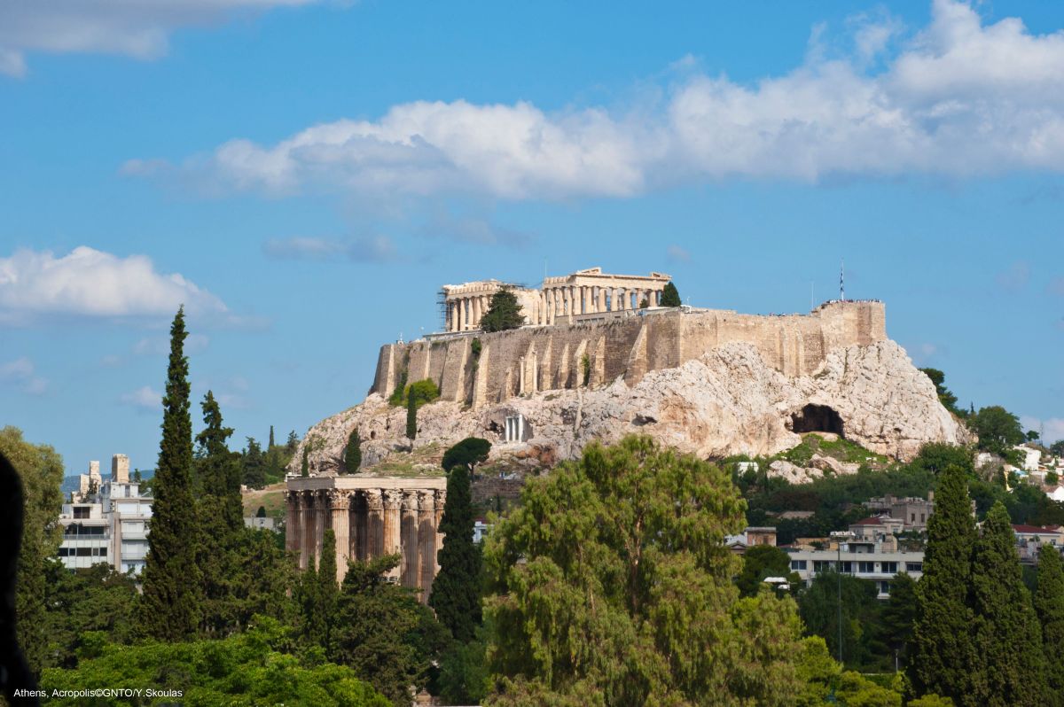 The ancient acropolis of athens, sitting atop a rocky hill with the parthenon temple and the temple of olympian zeus surrounded by lush green trees under a bright blue sky.
