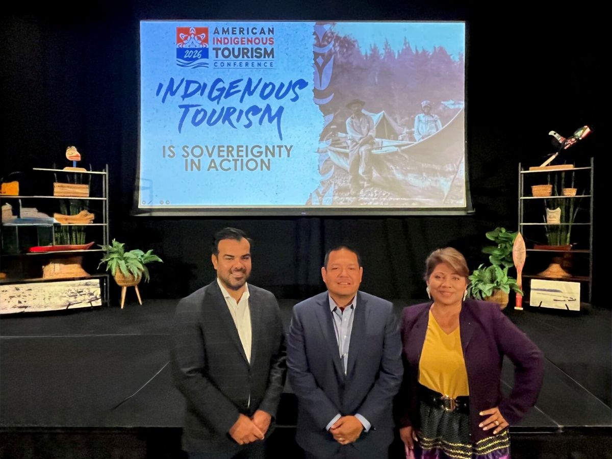 Indigenous tourism - travel and tour world Three speakers standing on stage in front of a presentation screen reading "american indigenous tourism conference 2026: indigenous tourism is sovereignty in action. "