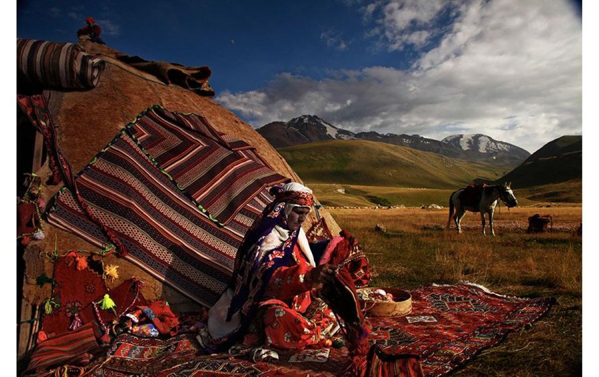 Iran - travel and tour world An iranian nomadic woman wearing traditional red and patterned clothing sits on a vibrant woven carpet outdoors, carefully crafting a textile. In the background stands a dome-shaped tent covered with striped woven blankets, set against rolling green hills and snow-capped mountains under a dynamic cloudy sky, with a saddled horse nearby.