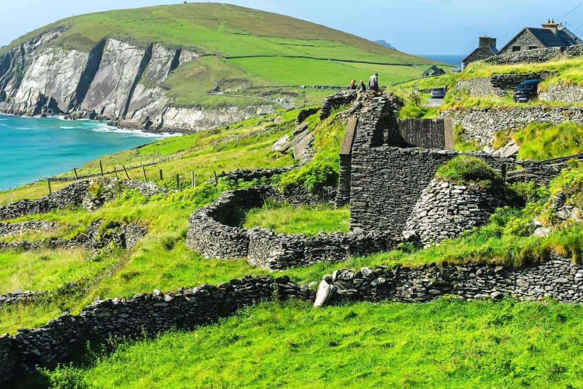 A scenic coastal view in ireland featuring lush green rolling hills, rugged stone ruins, and terraced stone walls overlooking turquoise blue coastal waters under a clear sky.