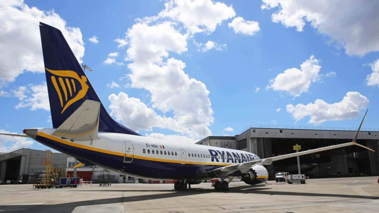 Ryanair aircraft parked beside airport maintenance hangars under a blue sky with white clouds.