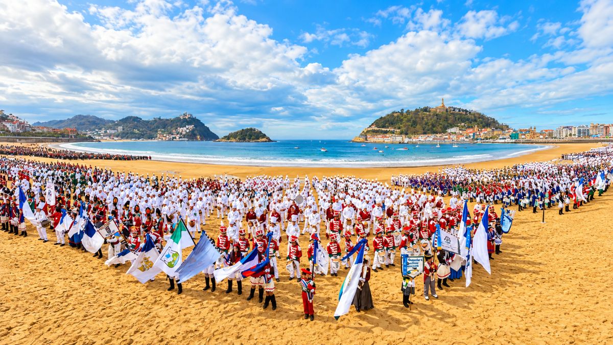 Large cultural parade on a golden beach with hundreds of participants in colourful ceremonial uniforms, flags and banners, blue sea, coastal hills, and a bright partly cloudy sky.