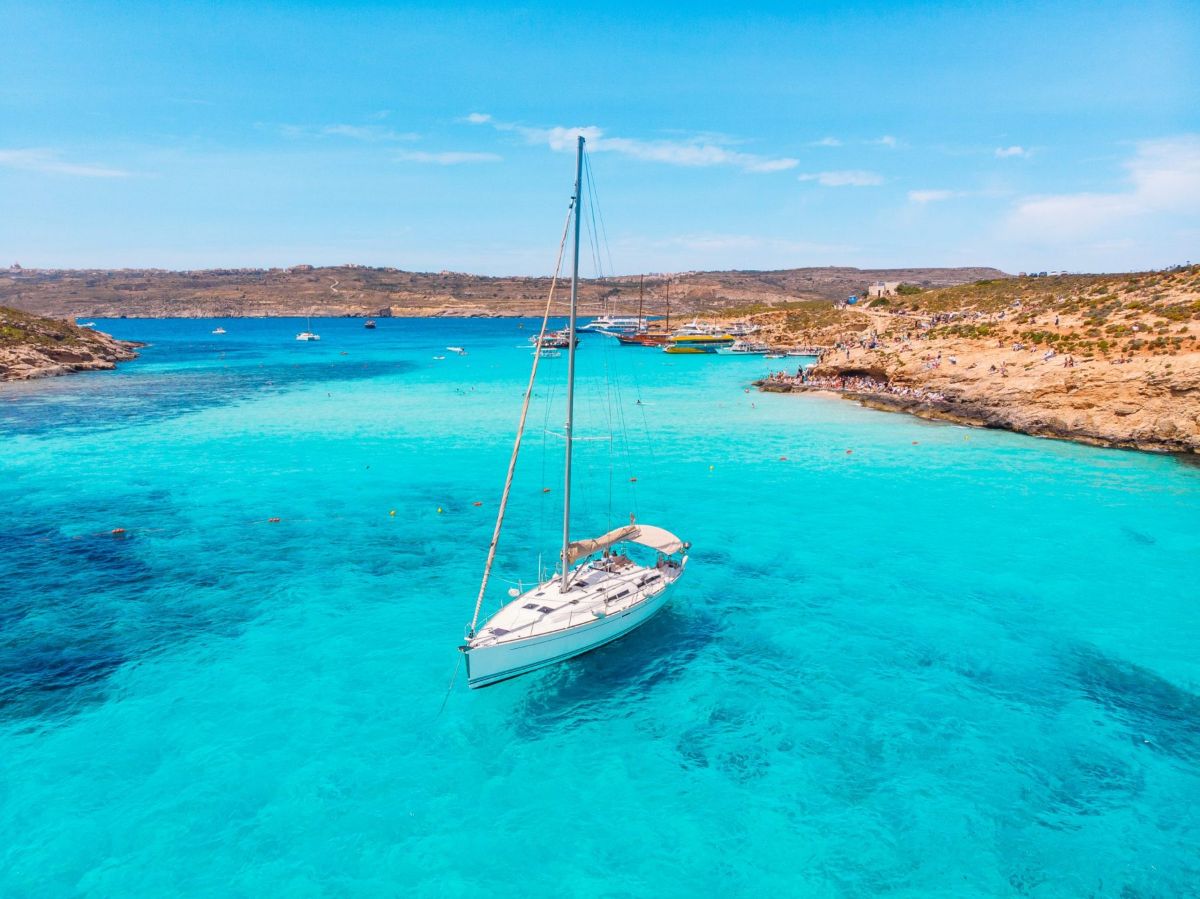 Malta - travel and tour world A white sailboat anchored in the crystal-clear turquoise water of the blue lagoon in malta, with rocky coastlines in the background.