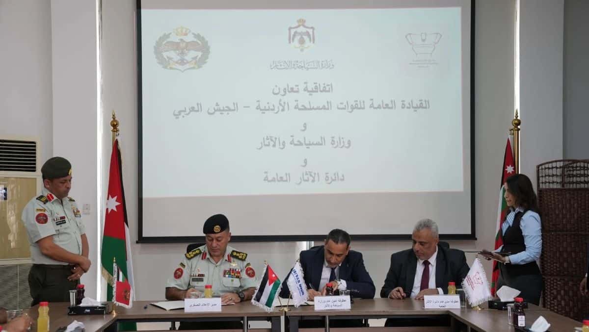 Three officials from the jordan armed forces &ndash; the arab army, ministry of tourism and antiquities, and department of antiquities &ndash; sit at a table signing a cooperation agreement in front of a presentation screen displaying arabic text and official emblems, with jordanian flags flanking the table.