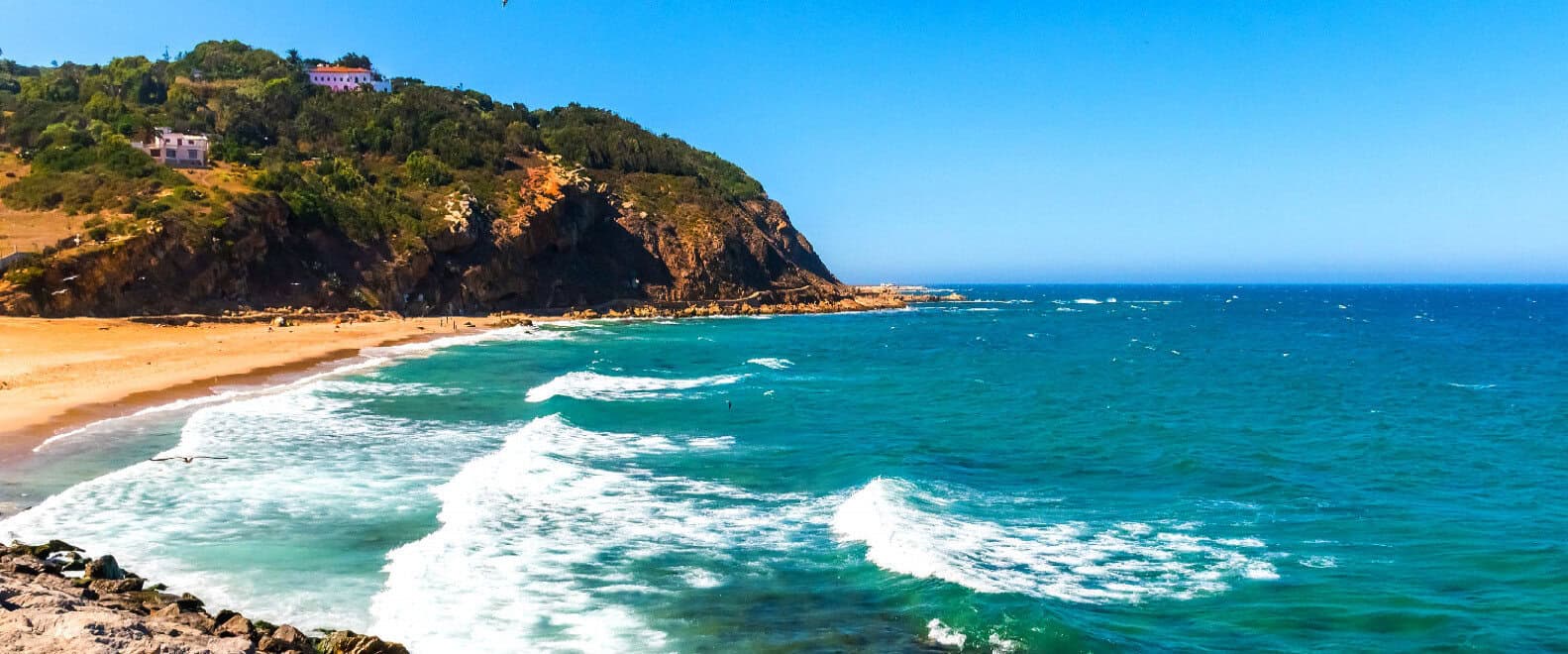 Turquoise sea and sandy beach below a green coastal headland in morocco.