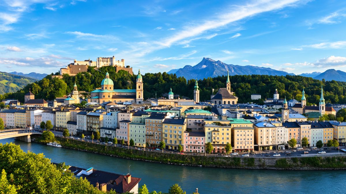 Panoramic view of salzburg&rsquo;s historic cityscape along the river, with pastel buildings, church domes, hohensalzburg fortress and the alps under a bright blue sky.