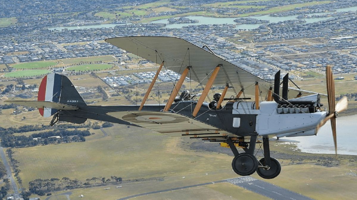 An aerial view of a vintage biplane in flight over a suburban and coastal landscape, showcasing its wooden structural struts, open cockpits, dual propellers, and a traditional tailfin marked with red, white, and blue stripes.