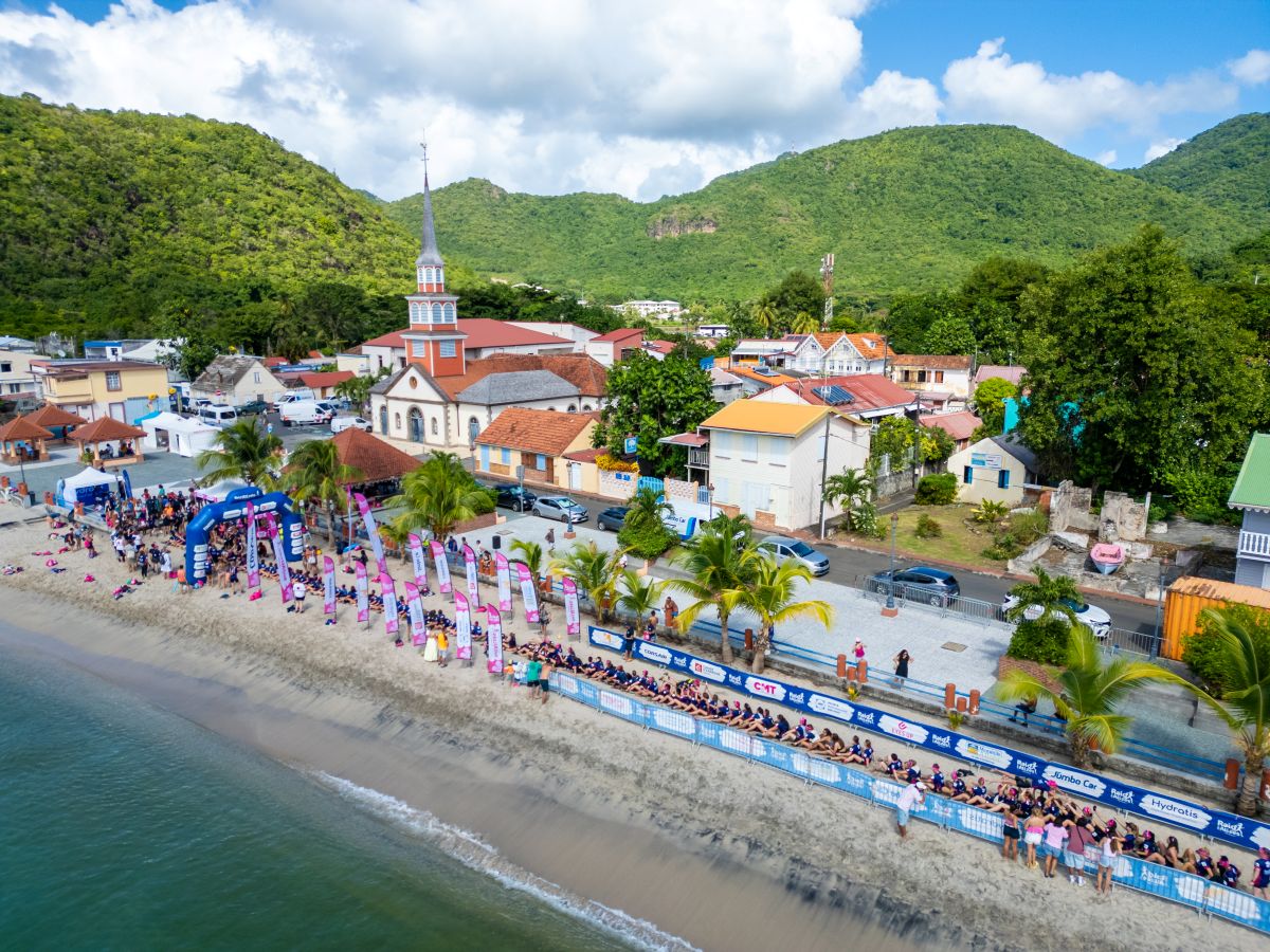 Raid des alizés - travel and tour world Aerial view of the raid des alizés event area on a sandy beach in martinique, featuring event arches, banners, participants, and a coastal village with a church set against lush green mountains.
