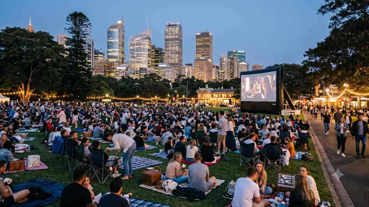 Saturday hotel economy - travel and tour world A large crowd of people sits on picnic blankets and chairs in a park at dusk, watching a movie on a giant outdoor screen with the illuminated sydney skyline and busy pedestrian walkways in the background.