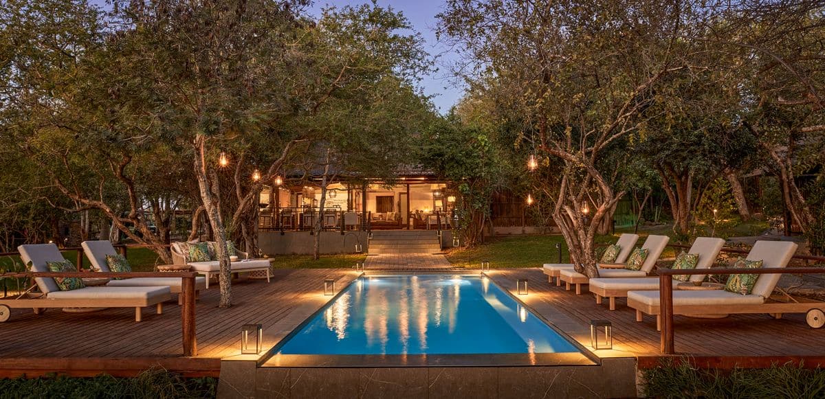 Illuminated swimming pool surrounded by wooden decking, cushioned sun loungers and trees at taj bush lodge